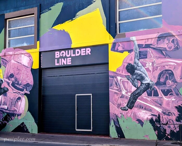 Homme en pleine action d’escalade sur une fresque murale très colorée, mêlant blocs de voitures stylisées et aplats de rose, violet, jaune et vert autour de l’entrée Boulder Line Avenue de Toulouse à Montpellier