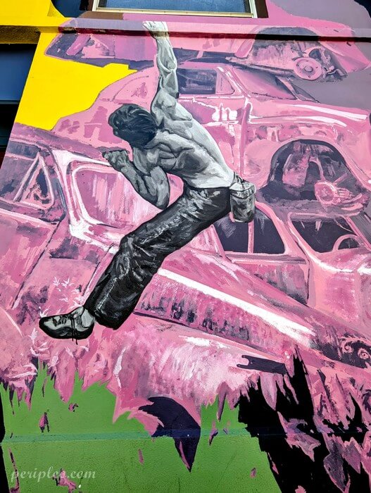 Street art autour de l’entrée Boulder Line Avenue de Toulouse à Montpellier, fresque monumentale mêlant hyperréalisme et univers industriel, représentant un homme suspendu à une corde devant un décor de carcasses de voitures aux teintes roses