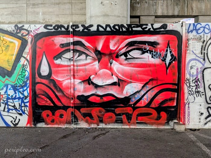 Street art sous le Pont de Sète à Montpellier avec grand graffiti rouge représentant un visage urbain dans une ambiance underground