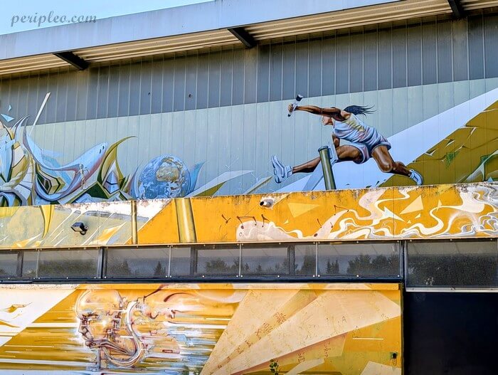 Fresque murale de street art sur un mur de la Halle des Sports La Condamine, représentant une athlète en plein saut de haies dans un stade, avec un style réaliste dynamique et des motifs abstraits dorés et blancs en arrière-plan