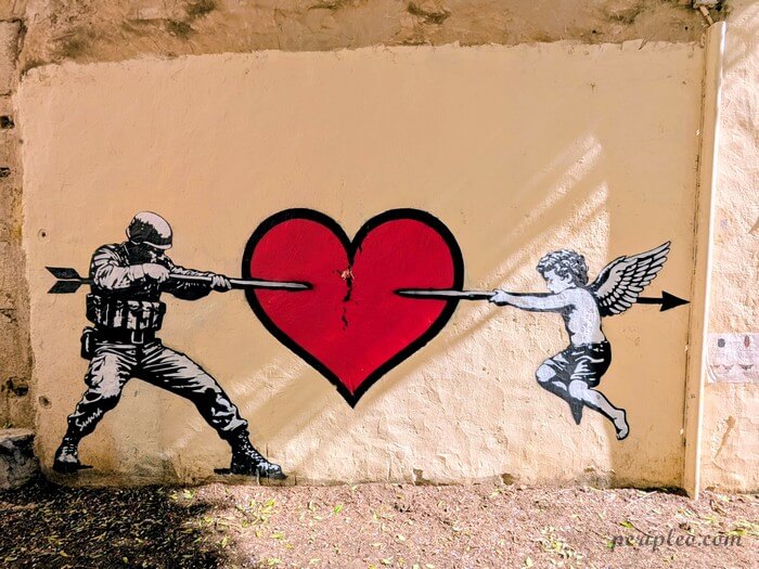 Street art Rue Saint-Pierre à Montpellier, œuvre engagée de Sunra représentant un soldat et un angelot se faisant face, chacun tirant une flèche à travers un cœur rouge fissuré