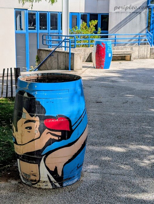 Baril peint de street art représentant un visage stylisé tenant un cœur rouge, dans la cour du Château d’Alco à Montpellier, avec un second baril décoré visible en arrière-plan, signé Sunra