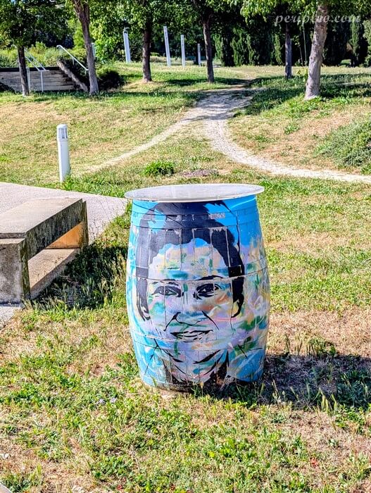 Baril peint par Sunra dans un parc du Château d’Alco à Montpellier, décoré d’un portrait stylisé dans des tons bleus et verts, posé sur une pelouse avec des arbres