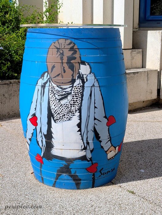Baril peint en bleu dans une cour du Château d'Alco à Montpellier, décoré d’une œuvre de street art signée Sunra représentant une silhouette au visage masqué, vêtue d’un foulard et d’un sweat gris, avec des cœurs rouges dispersés autour