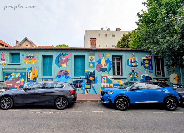 Fresque murale colorée de l'Atelier Parcours composée de formes géométriques et abstraites (visages stylisés, motifs et symboles) sur une façade bleu-vert, avec des voitures stationnées au premier plan dans la Rue Marcel de Serres à Montpellier