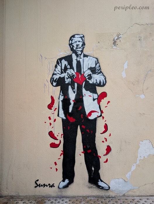 Graffiti représentant Donald Trump en noir et blanc sur un mur beige, debout en costume, en train de déchirer un cœur rouge dont des fragments tombent autour de lui, dans un style street art engagé signé Sunra